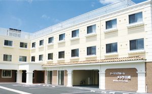 介護施設の建物外観。クリーム色の3階建て施設で、玄関ポーチとエントランス部分が見える。