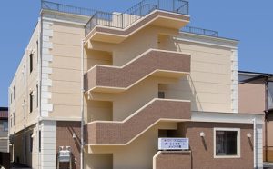 介護施設の建物外観。4階建てのベージュと茶色のモダン建築で、バルコニーが特徴。